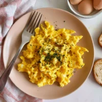 Fluffy creamy scrambled eggs served on a plate, topped with fresh herbs.